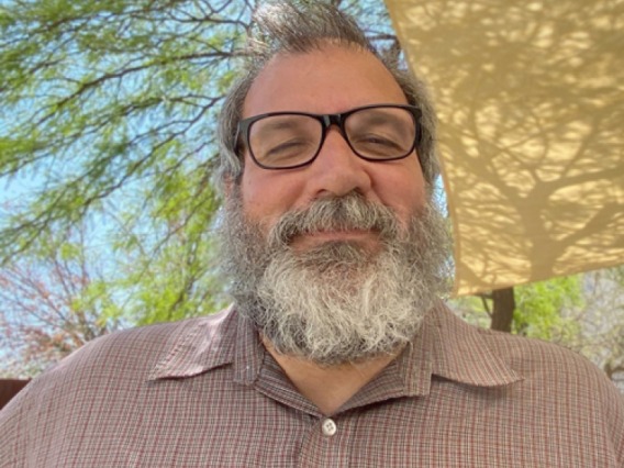 photo of JP wearing a tan pink shirt with black framed glasses and a grey/white beard