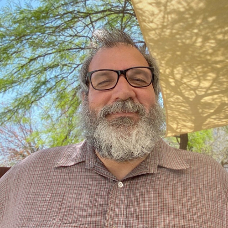 photo of JP wearing a tan pink shirt with black framed glasses and a grey/white beard